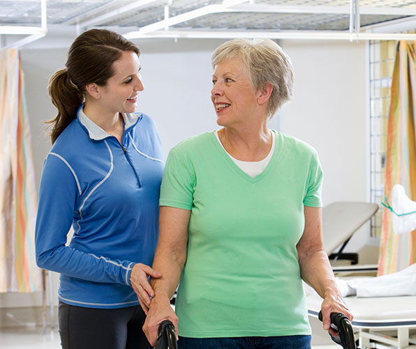 Young dark-haired female therapist standing next to an older female patient using a walker.