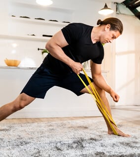 Man in a lunge position using a resistance band held under his foot to do tricep pulls.