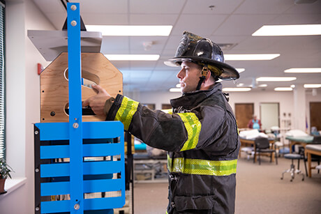 Male firefighter in full fire gear practicing safe lifting in a therapy gym.